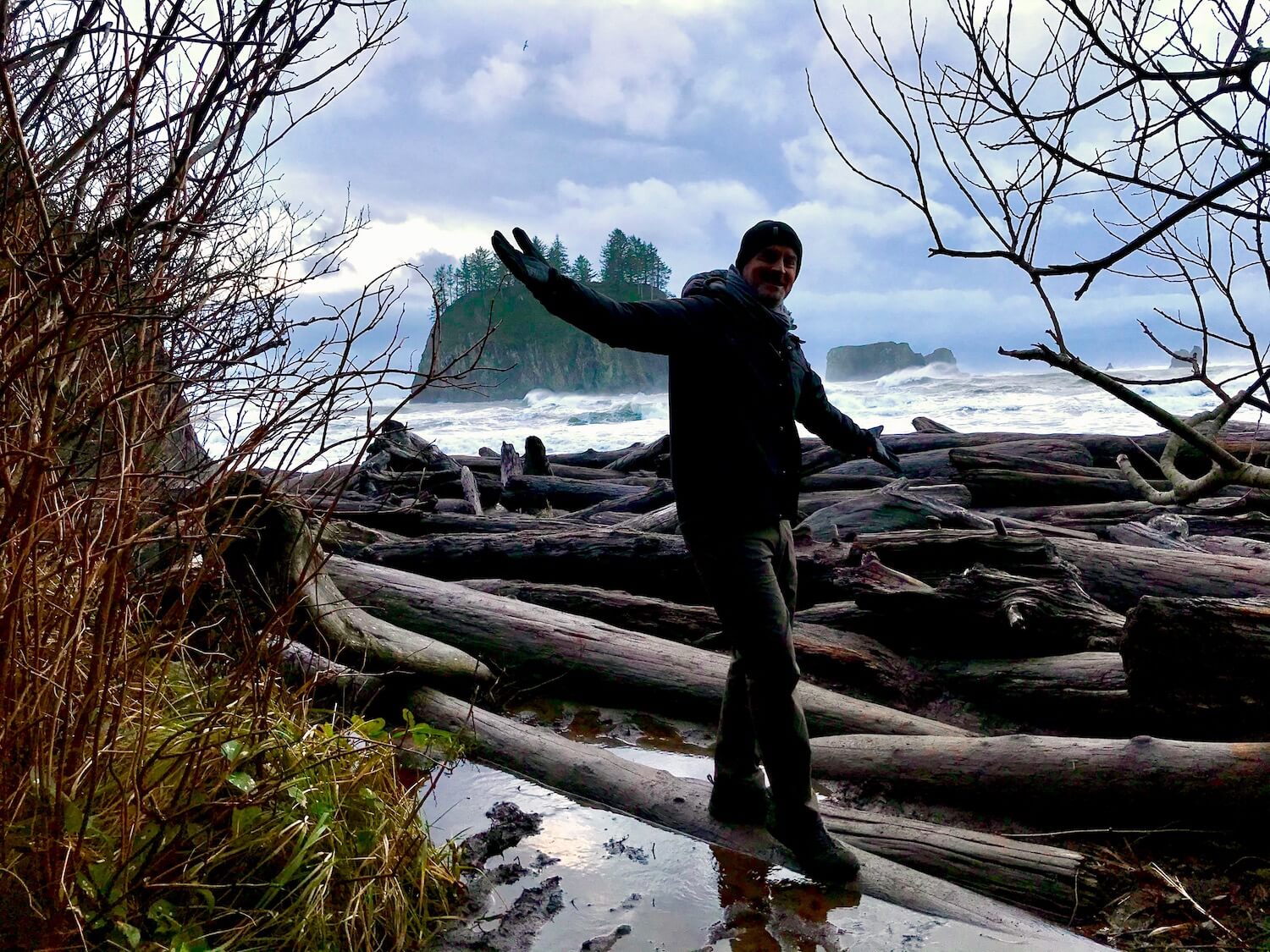 A man is walking on a log while exploring Second Beach in the Olympic National Park. The coastal waves are rough, crashing up against a barricade of wild logs, with a rocky island popping up in the background under a mix of clouds and blue sky.
