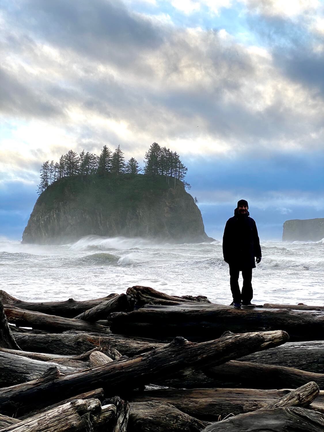 A beach scene of the Pacific Ocean waves crashing on a barricade of drift logs with a man dressed in black standing on one of the logs. A large rocky island in the background has fir trees growing on top. The sky is blue with a swirl of angry clouds.