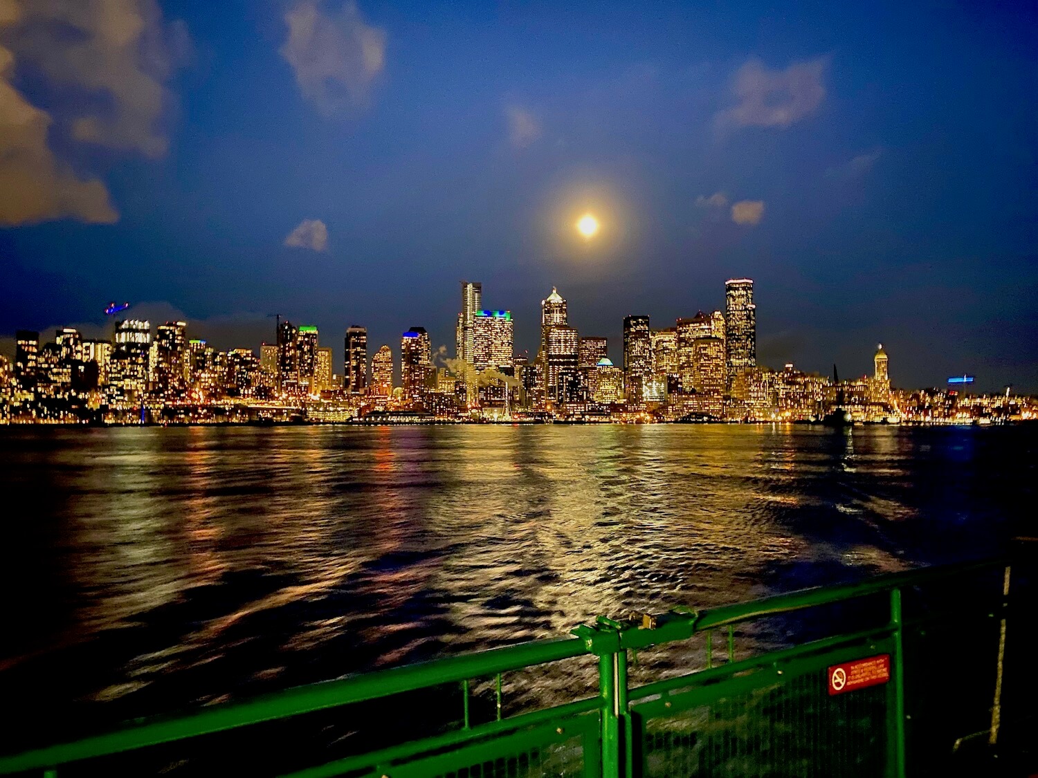 A view from a WA State ferry toward the lights of Seattle skyline. The moon shines above the buildings and blue sky with clouds in the dusk sky.