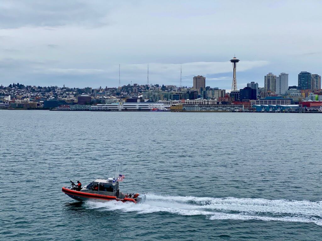 A coast guard speedboat follows the ferry with the Puget Sound in the foreground and the skyline of Seattle, with the Space Needle hovering above the buildings of the city.