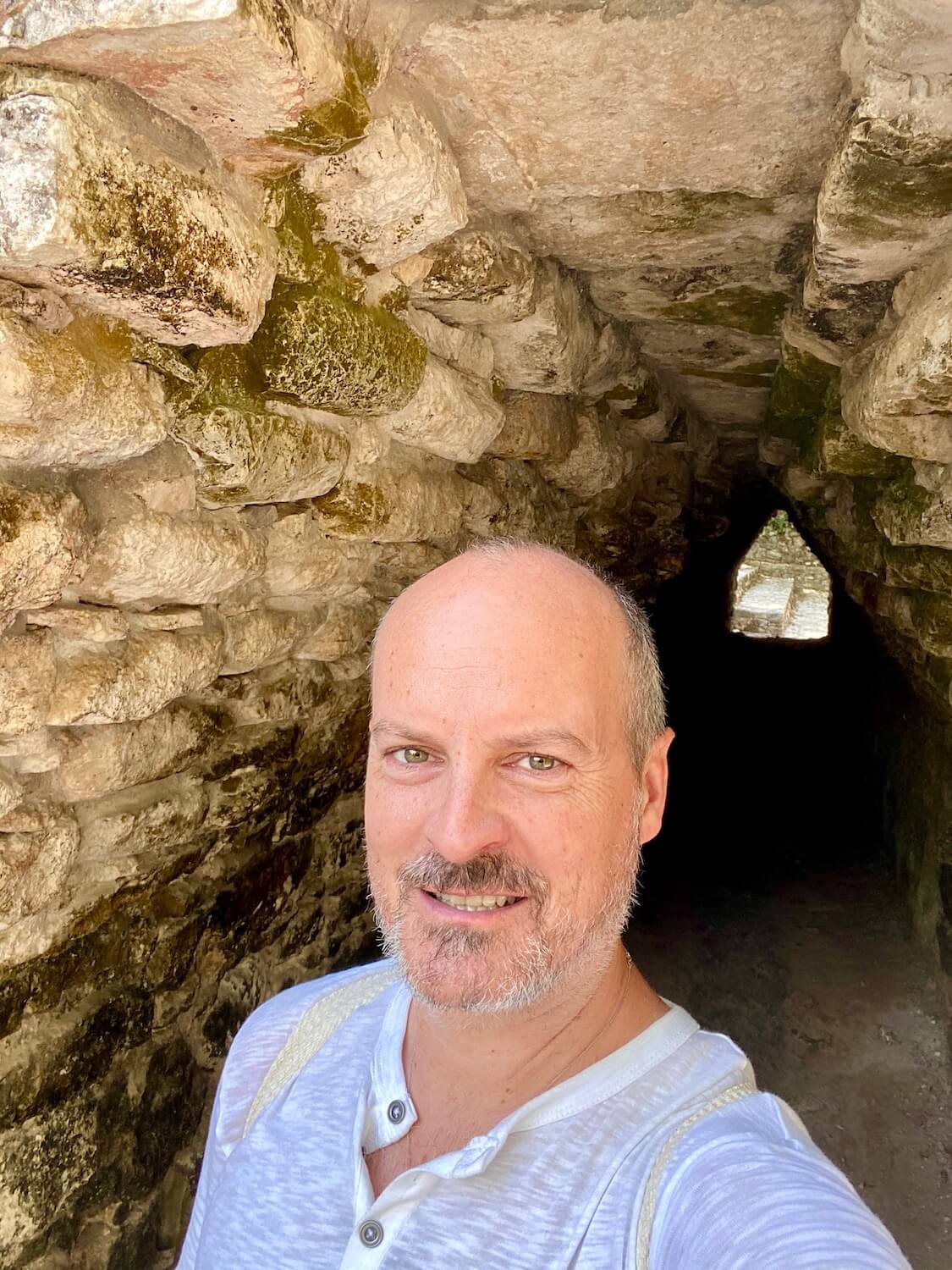Matthew Kessi at the foot of a fifty foot tunnel leading under the main stairs of the church pyramid structure in the Coba Ruins on the Yucatan Peninsula. He is wearing a white mesh t-shirt with beige straps from a sisal backpack.