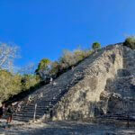 A wide view of 137 feet tall Nohoch Mul pyramid amongst the ancient mayan ruins of Coba. The mound of rocks is the tallest pyramid on the Yucatan Peninsula. Climbers make their way up and down the rock structure, many holding on to a rope affixed to the rocks for support. Jungle trees stand in the background amongst a bright blue sky day.