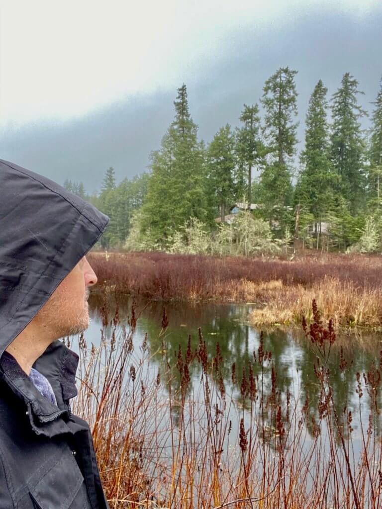 Selfie of Matthew Kessi looking out at Silver Lake. A black raincoat covers his eyes and the lake is bordered by red brush and tall fir trees further back. Silver Lake is located along Interstate 5 on the drive between Seattle and Portland.