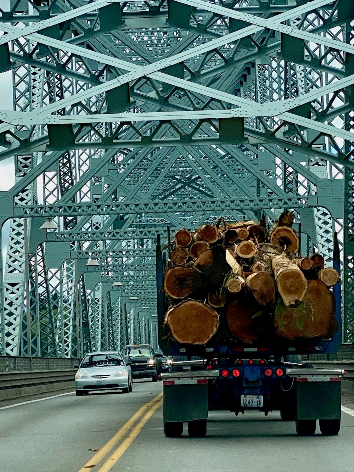 A logging truck full with wood heads up a bridge on the drive to Portland from Seattle on a bridge with aqua green erector beams supporting the entire structure. A car passes on the other side of the ride, which is divided by two solid yellow lines.