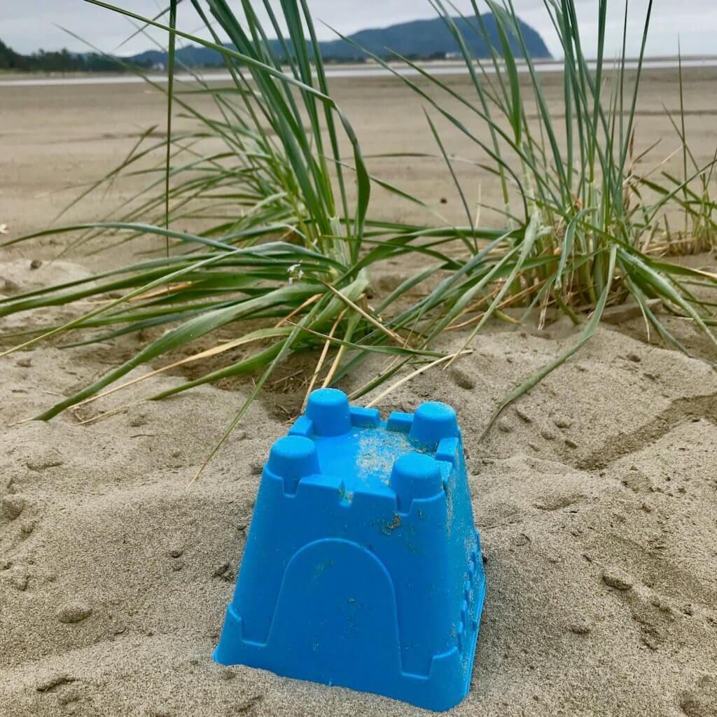 A single blue mold for a sand castle sits on a sandy beach on the Oregon Coast amongst wily green beach grasses, with the jetting outline of Tillamook Head in the background.