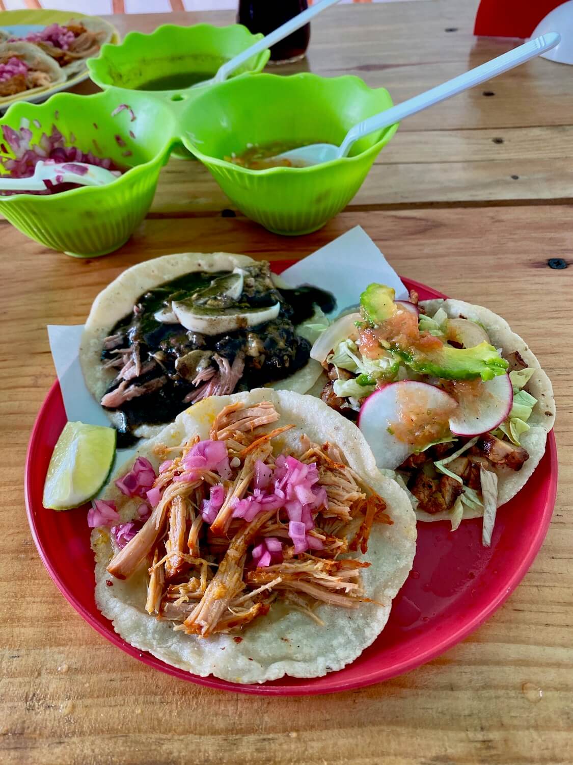A wood table with a tasty looking plate of Mexican tacos is accompanied with three plastic green bowls with onions, green Salsa and spicy pico de Gallo and white spoons in each bowl.  The tacos are served on a red plastic plate and are three unique varieties.  One taco has pork with a black bean sauce another pork with radishes and avocado and the closest taco to the camera is pulled pork with a sprinkling of red onion on top.  There is a lime wedge as garnish.  