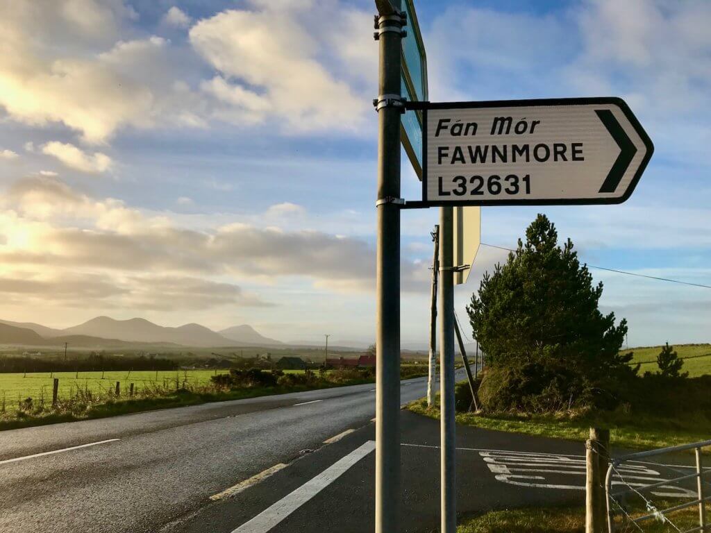 A road sign along a main route in County Donegal in Ireland.  The sign says Fan Mor in Irish and Fawnmore in English and has a black arrow pointing to the area off the road.  In the background are rolling green hills and taller mountains in the background with blue sky peeking through fluffy grayish sunset clouds.  