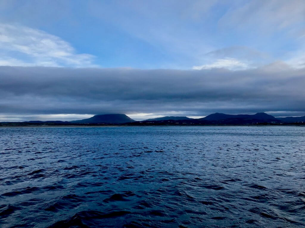 The icy blue North Atlantic Ocean from a ferry boat crossing between Tory Island and the mainland Donegal in Ireland.  The waves are rolling as a cloud layer in the distance slowly sneaks away from the mountains in the background, including just the cap of Muckish Mountain.  The sky above is already a vibrant shade of light blue. 
