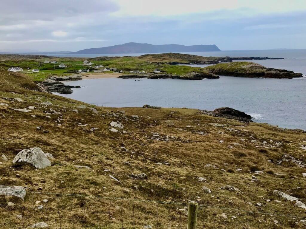 A view of the Irish coastline in County Donegal.  The landscape leading down to the sea is brownish yellow and depicts very rocky, inhospitable soil with a stretch of sandy beach along a quiet cove.  Near the beach is a grouping of ten homes, barns and other smaller supply buildings.  In the far distance is the commanding peninsula of Horn Head, jutting out into the ocean.  Blue sky above is beginning to be over taken by grayish rain clouds coming from the left side of the photo.  