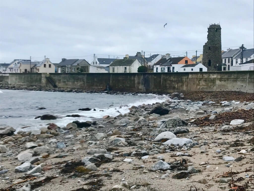 View of the rock laden beach near the ferry dock on wild Tory Island, the northern most island in Ireland.  The waves are crashing on the rocks, creating foamy white surf and the drab gray seawall protects the many houses seen in the background from the wild coastal waters.  In amongst the homes, with their concrete walls and slate roofs is a 4th century stone tower, still barely standing.  