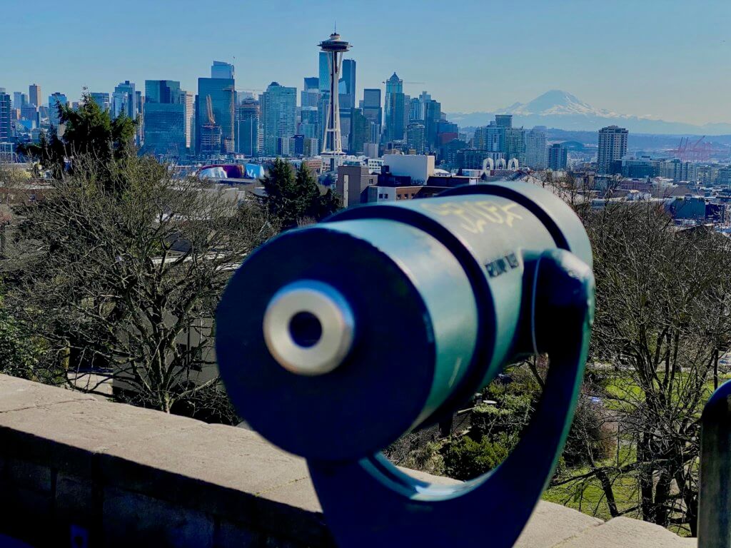 View of Seattle from Kerry Park on Queen Anne. In the forefront is a telescope facing towards Mt. Rainier in the background rising up above the skyline of the city of Seattle. The Space Needle graces the line of buildings in a prominent manner with the futuristic looking building. The sky is blue and the grass in the foreground amongst deciduous trees.