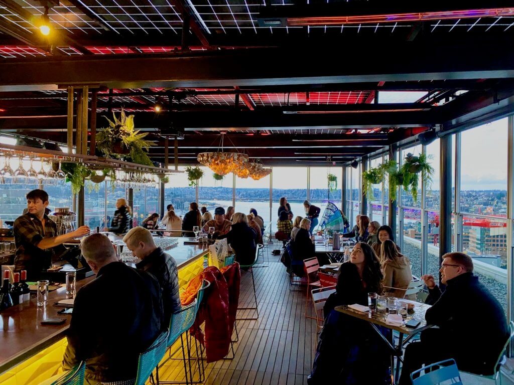 MBar in South Lake Union area of Seattle has a modern hip rooftop bar and restaurant and this photo shows a bartender mixing drinks with two guests sitting at the bar and other patrons scattered amongst tables in a section enclosed by glass windows. In the background is an open view of Lake Union, while the roof has grids with bright pink and purples neon lights glow.