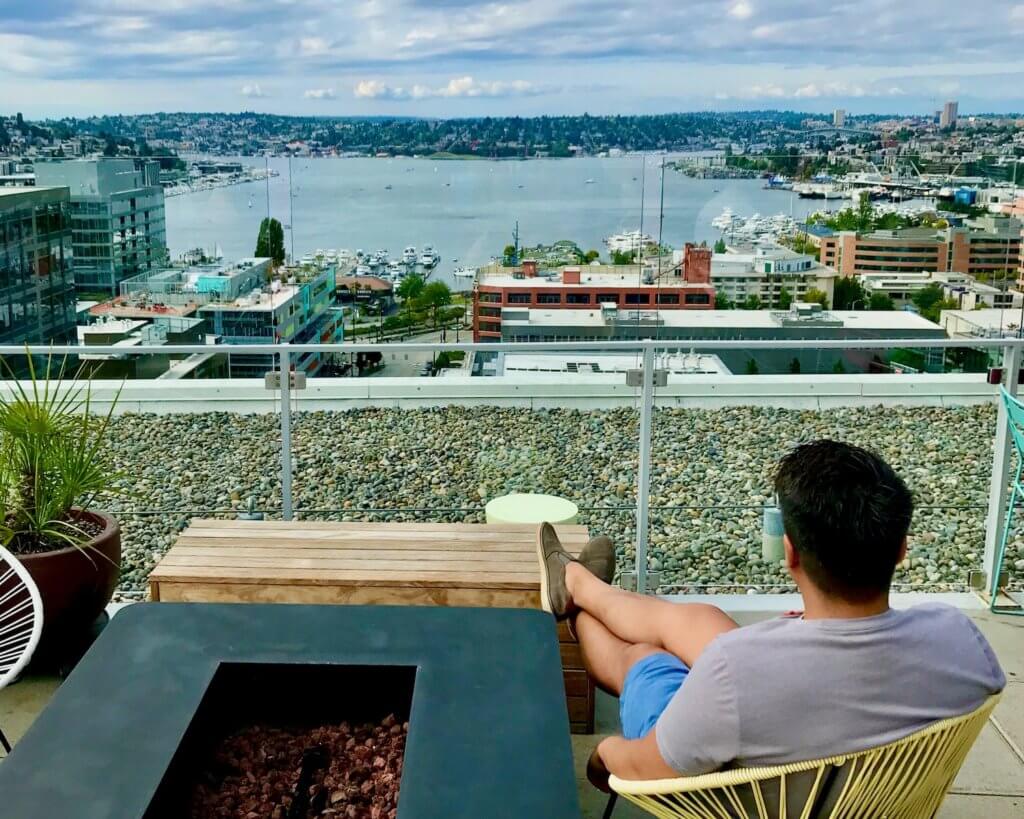 View from rooftop of M Bar looking out towards Lake Union. A guest sits in a lounge chair facing the view and the fireplace next to him is idle. The lake is blue and has boats under a mostly cloudy sky.