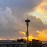 Sunset view of the Space Needle taken from the rooftop bar in South Lake Union area of Seattle. The clouds are dramatically gray and open up to a small patch of blue sky and other brightly burning hues of orange pink and purple in the winter sunset glow. In the foreground, darker office buildings frame in the base of the inconic tourist attraction.