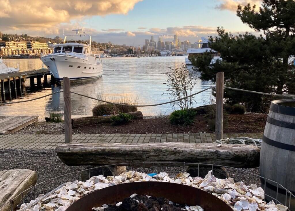 View from a bench next to a fire pit that is not in use. The fire pit is surrounded by oyster shells and in the background Lake Union leads to a faint skyline of all the tall buildings in Downtown Seattle. A yacht is moored at a dock leading from the restaurant area and a pine tree frames in the right side of the photo.