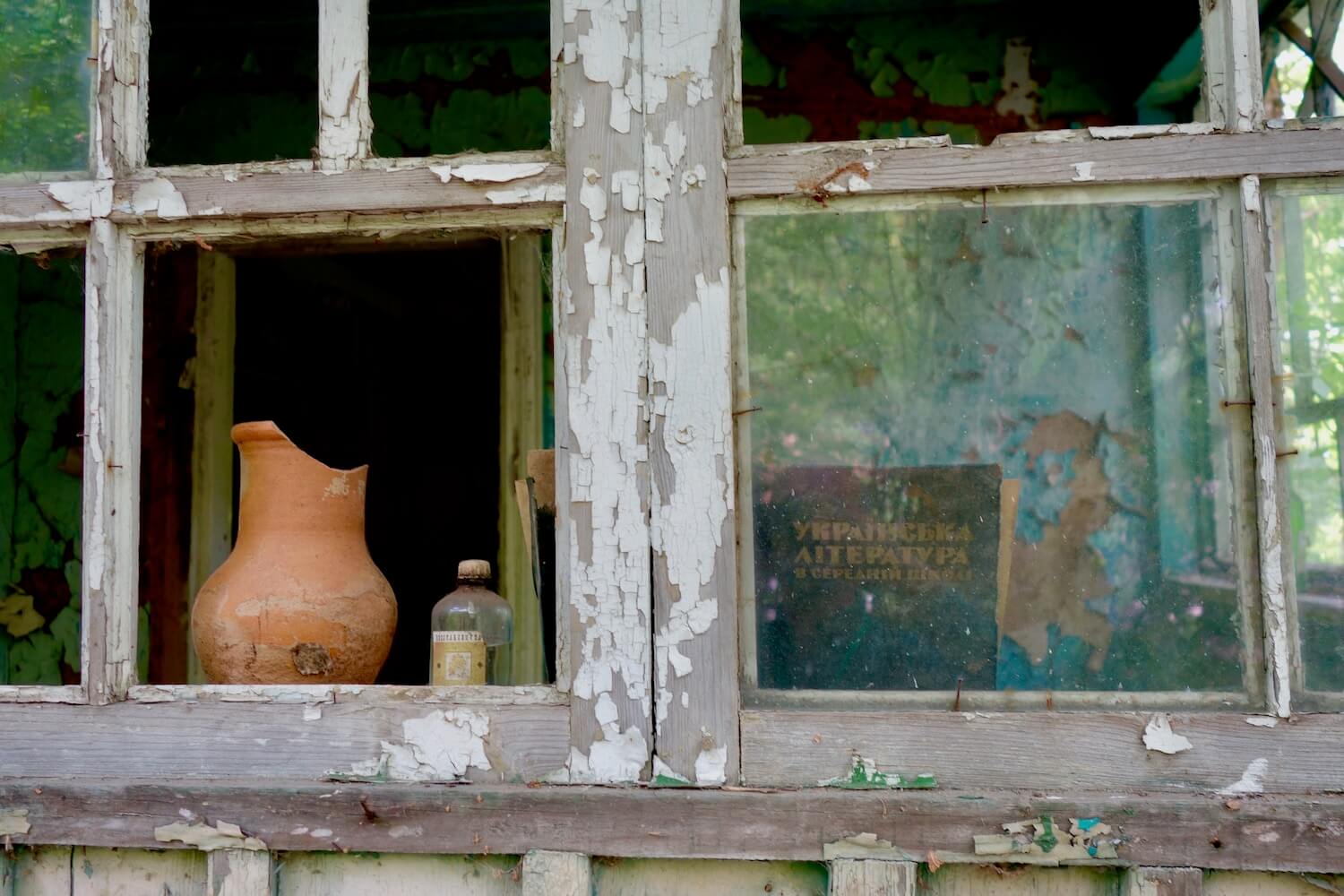 An abandoned home in the small village of Chernobyl in Ukraine.  This shot looks through a broken window with gray paint chips flaking off the frame.  Inside on the window sill is a clay picture with a chip on the top and some medicine in a glass bottle.  Behind another window pane is a book with Russian writing on the cover.  The inside room is an aqua color with the surfaces of the wall peeling away.  