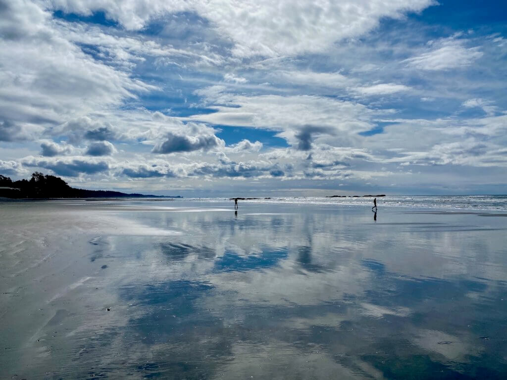 Broad shiny wet sand near Kalaloch Beach, with the waves of the Pacific Ocean crashing up on a low tide beach, wet with water and reflecting the swirl of blue sky with white puffy clouds. There are two figures walking on the beach in the distance.