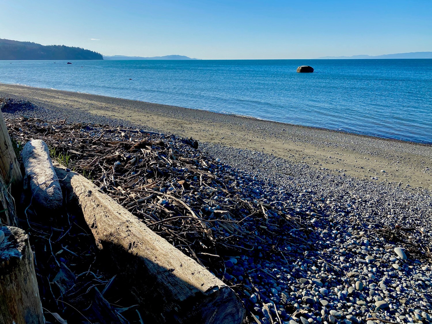 The view from charming Joyce Washington allows sights of both Vancouver Island in Canada and the tip of Washington State in this view of a pebbly beach with drift logs and sea debris leading to a peaceful blue body of water with slight ripples of waves.  The sky in the background is blue.  