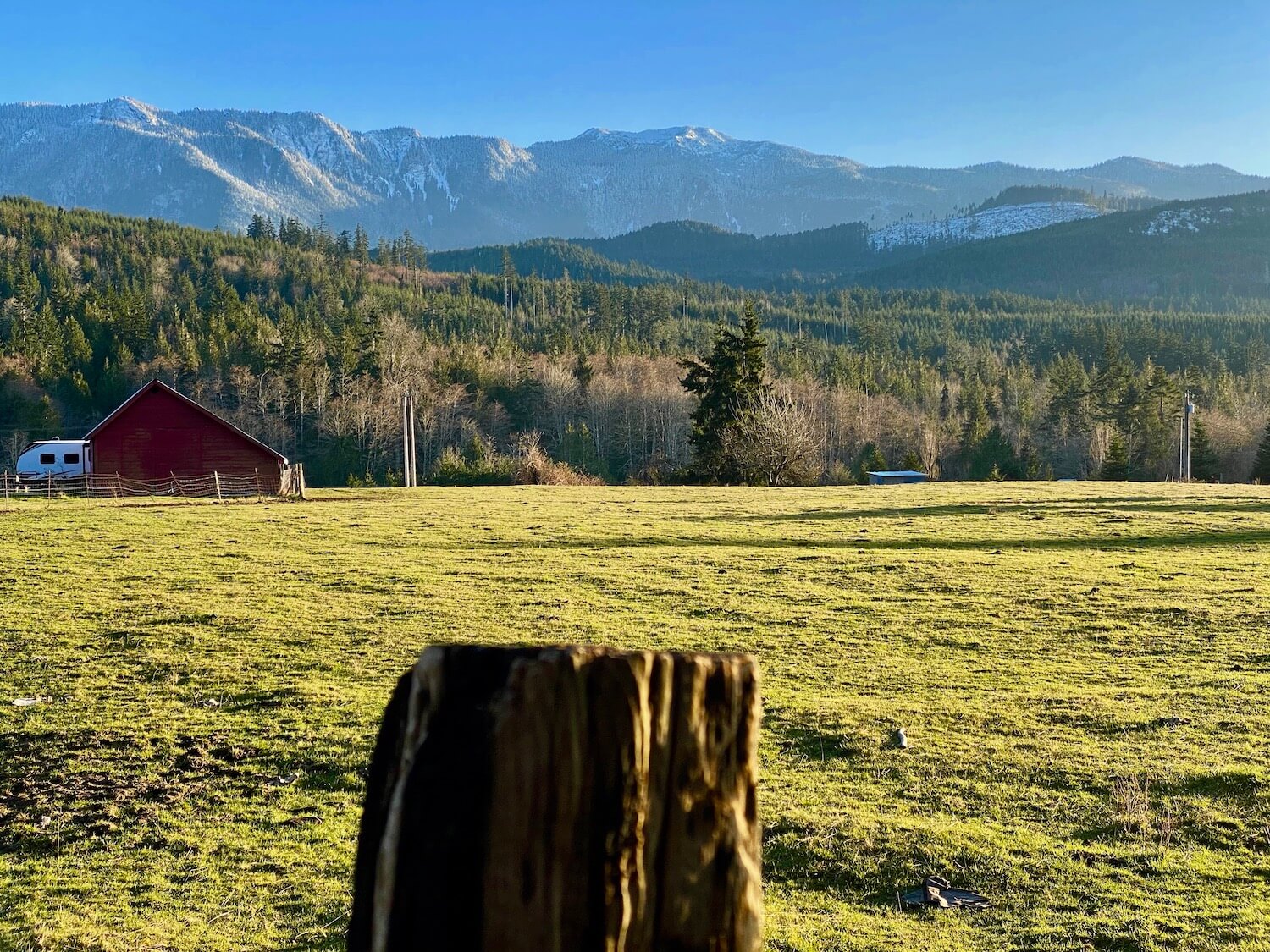 The rolling foothills near Joyce Washignton lead up to the lightly snow covered Olympic Mountains. In the foreground is an out of focus fence post and the pasture is bright lime green with a red barn in the distance.