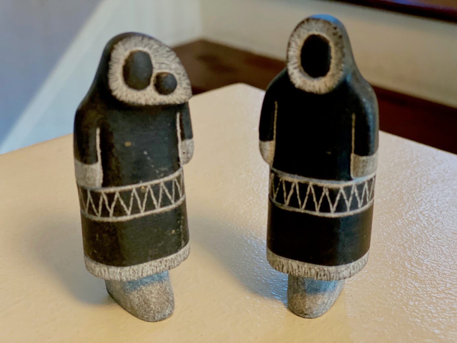 An inuit couple, man and woman with baby carved from soapstone. The carving is done by a local Greenlandic artist in a way that shows black covered outer coats with lighter animal fur fringe around the faces hands and feet areas. These two small figures come from Nuuk in Greenland.