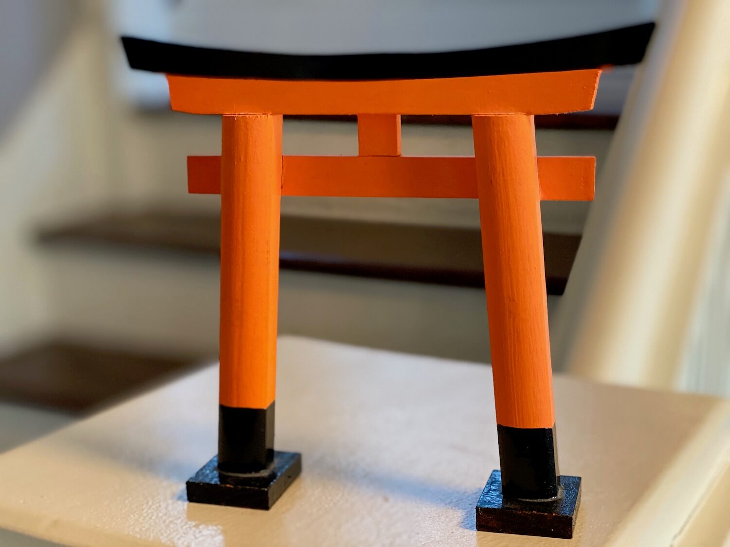 A miniature replica travel souvenir with soul, representing one of the hundreds of gates at the Fushimi Inari Shrine near Kyoto Japan. The gate is made up of two columns, each with a black lacquered base leading up to a orangish red pergola connecting the two. The Japanese style cap of the pergola is also black lacquer.