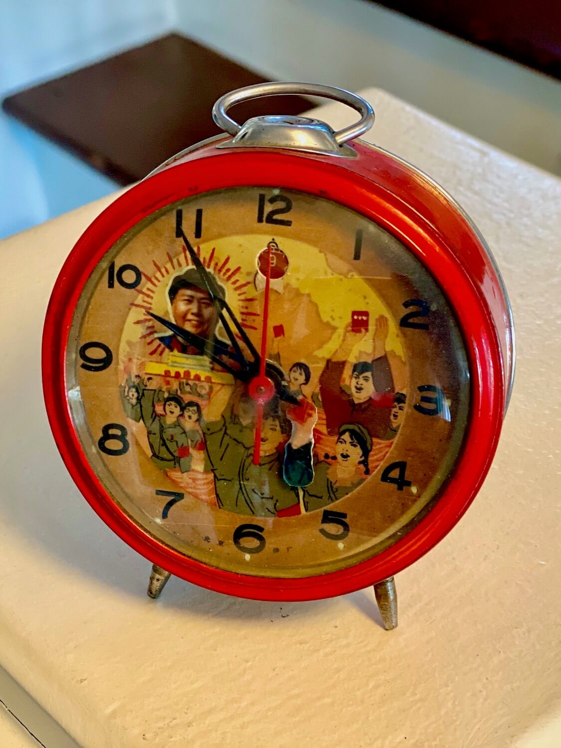 This alarm clock is vintage from 1950's era China and depicts Mao Se Tung and other communist characters inside the clock hands and western numbers. The body of the clock is bright Chinese red while the legs are aluminum color as well as the loop on top.