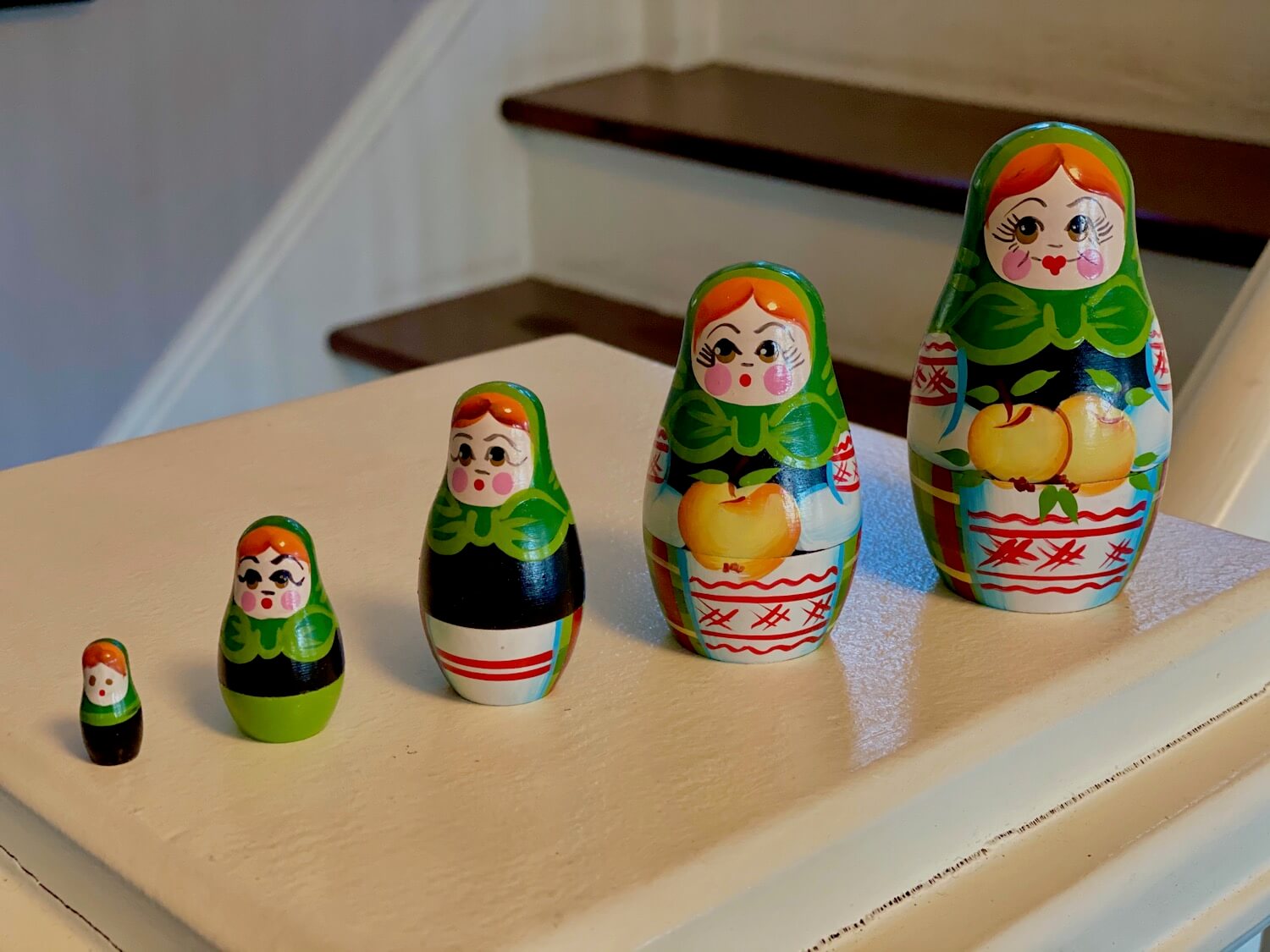 A set of five Russian style dolls purchased in Minsk, Belarus. Each doll gets progressively smaller. They are all depicting the same women wearing a green scarf, blond hair, black shirt while holding apples.