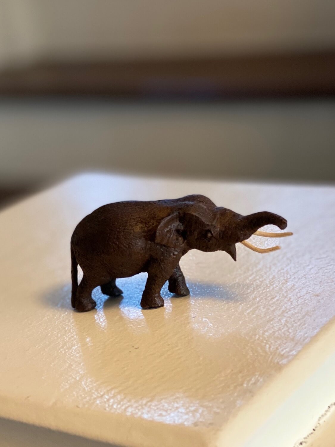 A miniature carved elephant made from dark brown mahogany. The elephant has two white tusks and is in a position as if calling out, with trunk protruding horizontally outward. This was purchased in the shop adjacent to the renown restaurant Bo.Lan in Bangkok Thailand