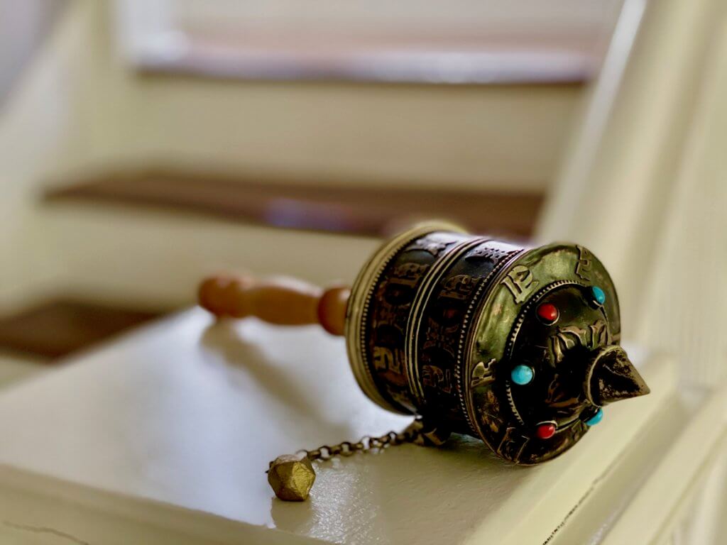 This prayer wheel comes from a Tibetan monastery in the Ladakh region of India. The round metal casing is a rubbed bronze color and a small bronze chain connects the main cavity of the prayers to a small ball that is used to twirl the wheel around. On the top of the wheel are decorative gems of red and aqua and the top has a pointed hat like nob.