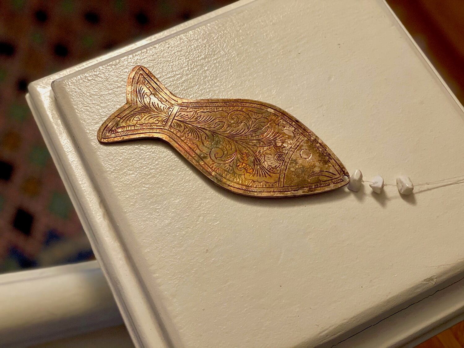 This silver fish is connected to fishing line and three small white stones. It comes from near the ancient site of Dougga near Tunis in Tunisia. In the background on the floor is a hand crafted Tunisia rug with many small diamond shaped figures of different colors.  Truly a souvenir with soul that reminds of great travels.