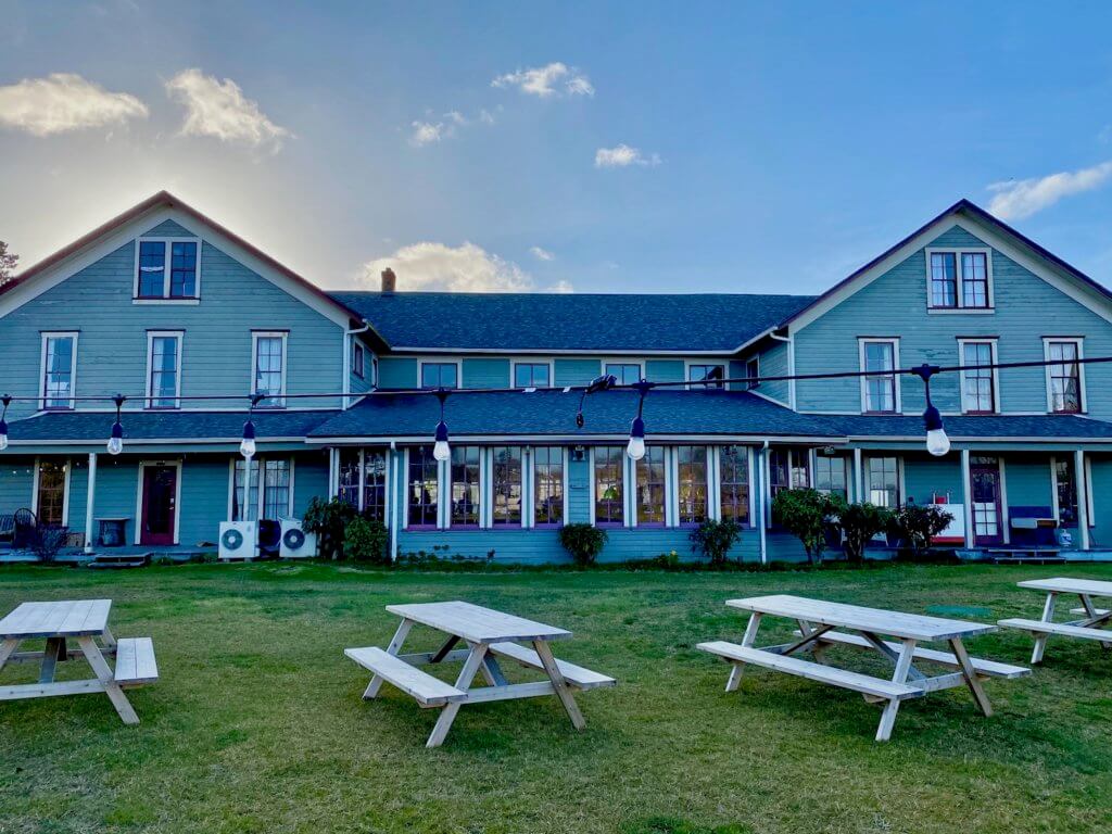 The front view of the Tokeland Hotel, which is Washignton State's oldest continuous operating hotel. The wood siding is a blueish green color and the windows on each of three stories have eight panes each with white trim. There is a strong of lights running across a gathering area on the lawn in front of the hotel with four rustic picnic tables. The sky is blue with white puffy clouds.