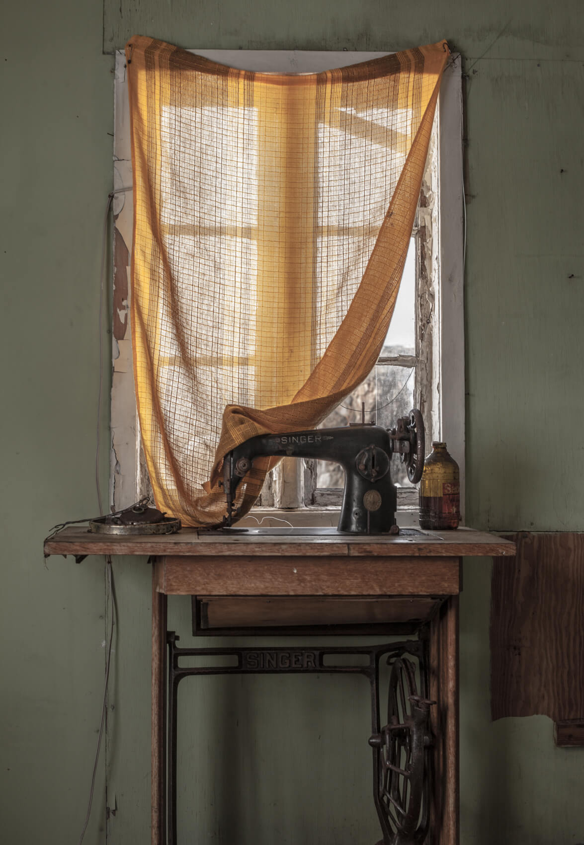 A room inside an abandoned home in the north of Greenland. An old sewing machine and table sit in front of a window covered by dingy orange see through curtains. The wall is pale green with pieces of plaster missing.
