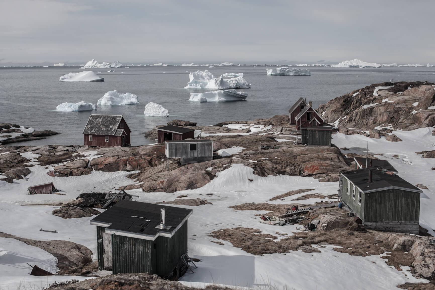 An abandoned village in Northern Greenland is uncovering from the snow, which is slowly melting away from the base of several of the houses, built on a rocky coastline to the Arctic Ocean, where huge chunks of ice are flowing in the icy gray waters. The houses are either red or gray and mostly one story high.