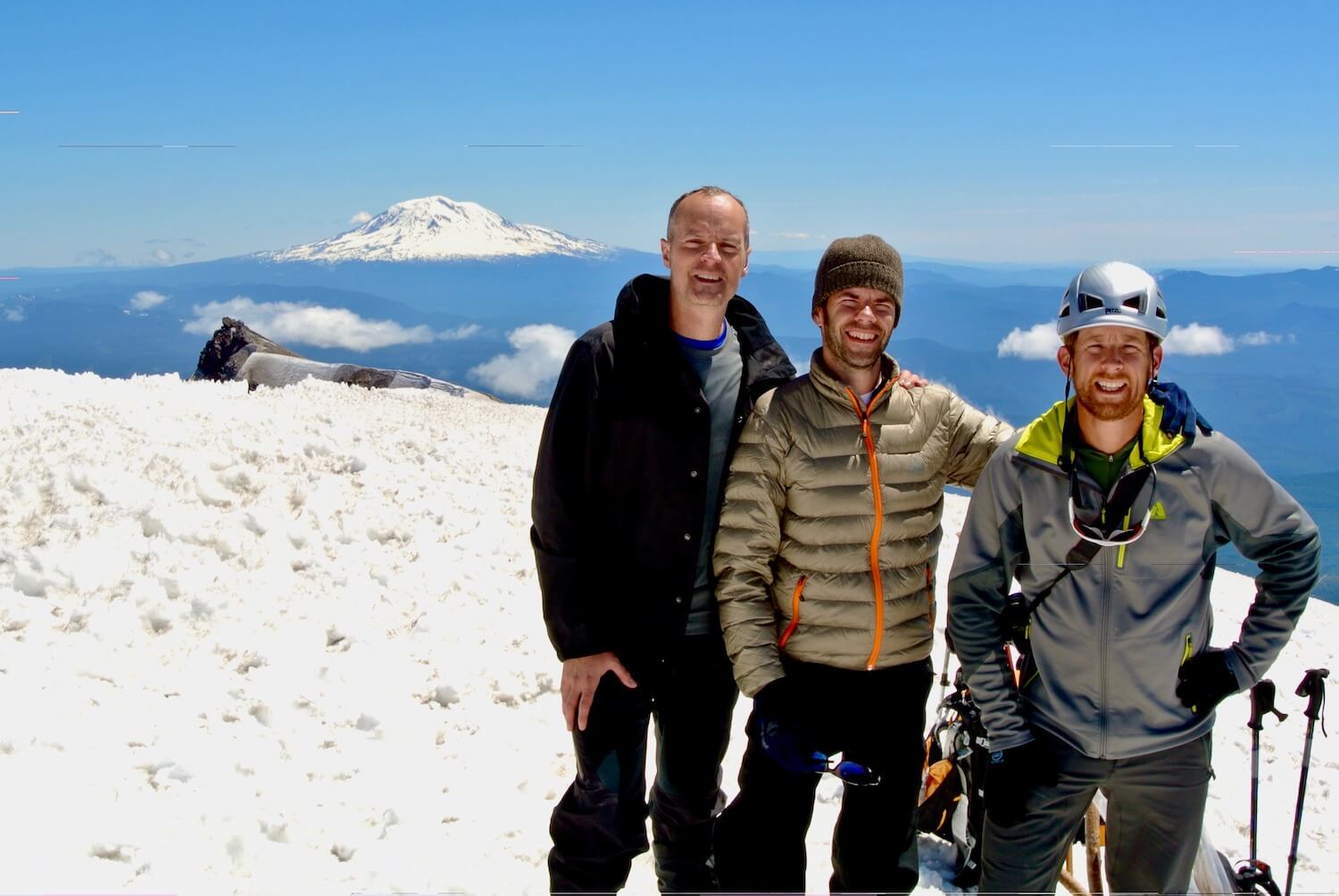 Three hikers at the summit of Mt. St. Helens, with the rim to the crater in behind them in gray. There is snow on the ground and they are wearing hiking gear. In the distance Mt. Adams rises up amongst blue skies.