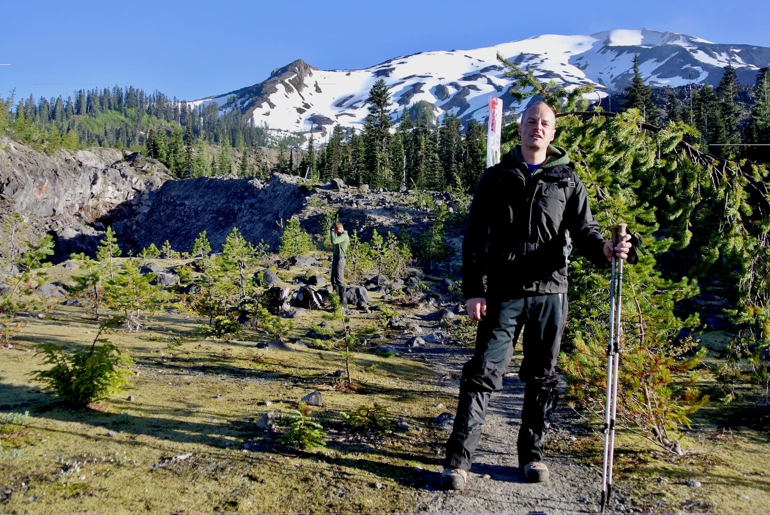 A photo of Matthew Kessi preparing to hike to the crater rim of Mount St. Helens.  He is holding hiking poles and wearing black waterproof gear.  The mountain is still mostly snow covered with patches of gray rock and in the foreground small fir trees pop up from a mossy open trail.  
