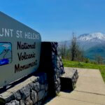 The entrance sign to the Mount St. Helens National Volcanic Monument. The sign is housed on a rock wall and sits on the side of a steep hill facing the snow covered volcano in the distance. The grass near the sign is bright green with the blue sky.