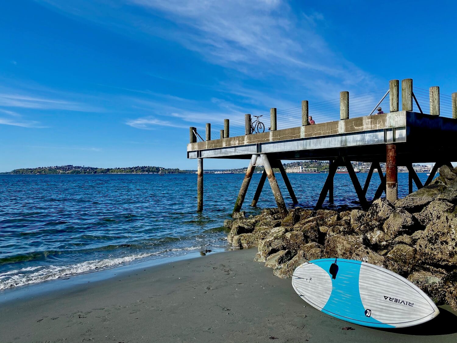 Stand up paddle boarding is a popular thing to do in West Seattle and this photo shows a blue and white paddle board leaned up against some large barnacle covered rocks supporting a metal and concrete pier which juts out into the Puget Sound from Alki Beach in West Seattle. The water is greenish blue and sky bright blue with a few scattered white clouds.