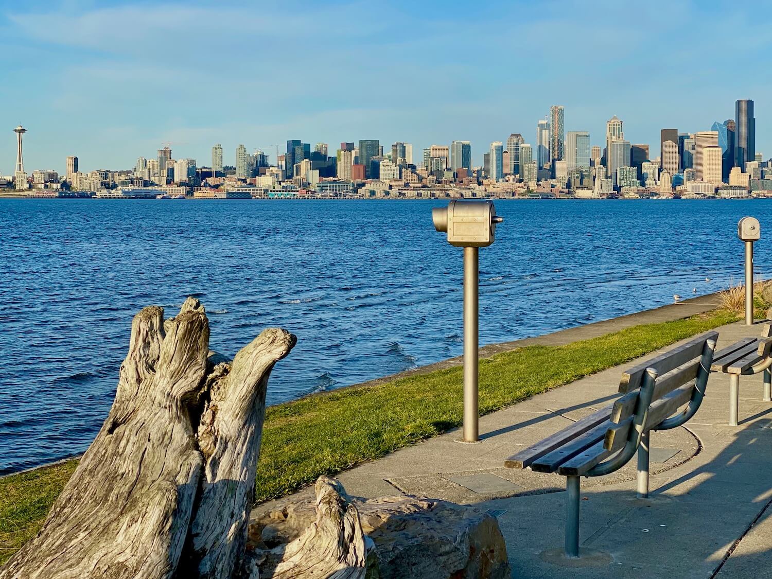 Alki Beach and West Seattle offer sweeping views of the city and this shot shows the skyline from the Space Needle to the tallest black building, Columbia Center. The city is across Eliot Bay, which is a rich blue color. In the foreground is a large piece of driftwood as well as two silver telescopes and two slatted benches.