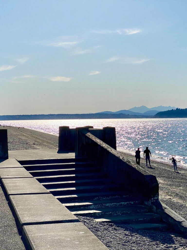 Walking Alki Beach is a great thing to do in West Seattle, especially during nice weather like in this photo. There is the silhouette of a couple and a child walking on the sandy beach near the edge of the water and in the foreground concrete steps lead right into the pebbly sand. The Olympic Mountains are on the horizon as well as Bainbridge Island as the late afternoon glow starts to set in with light blues and peach horizon.