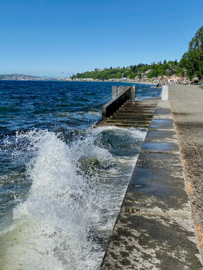 Waves crash onto the seawall at Alki Beach in West Seattle. The Seattle Space Needle is in the distant background under bright blue sky.