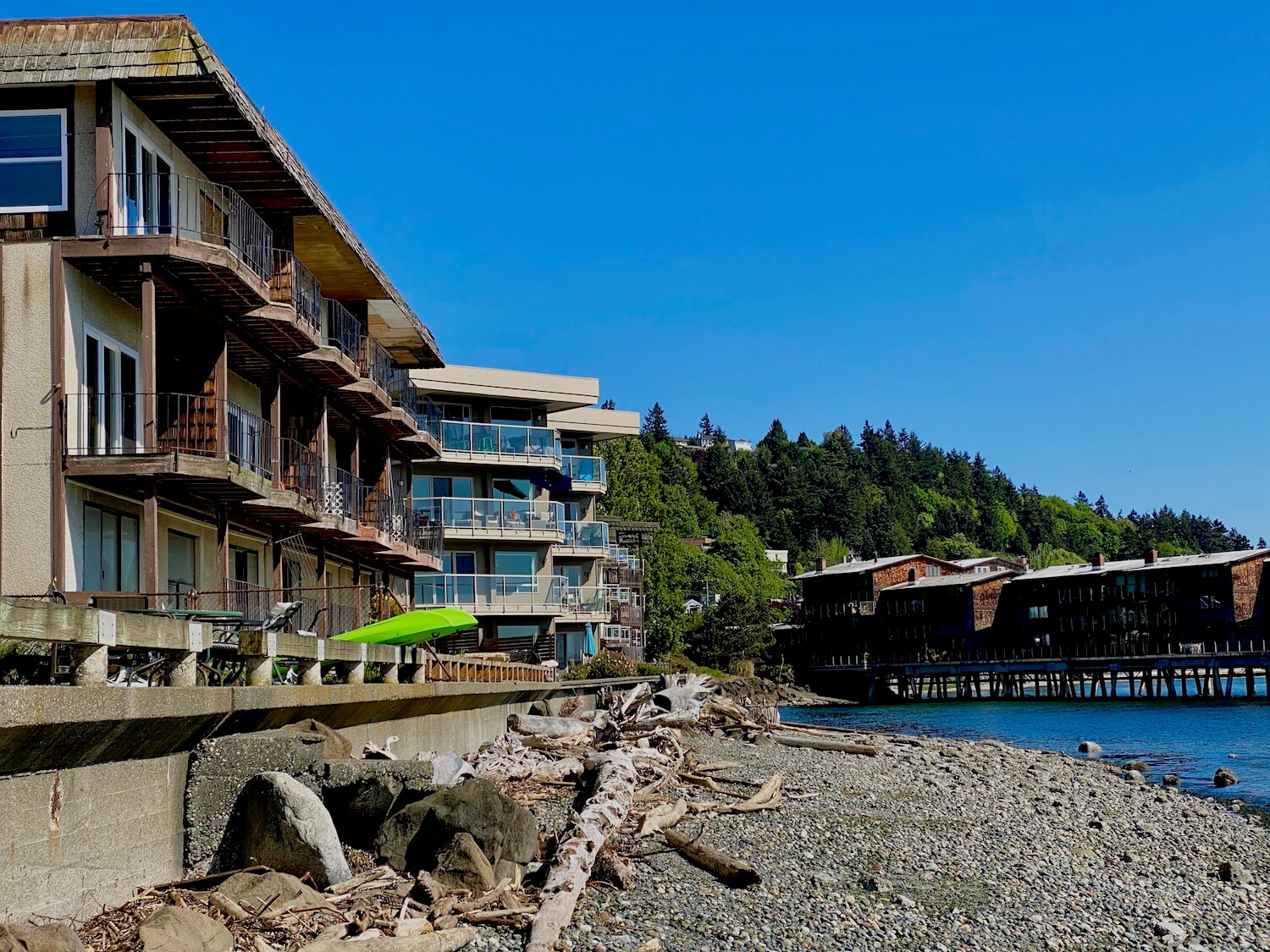 An apartment building on Alki Point in West Seattle appears to be going through renovations while two lime green kayaks hang out on the seawall. The beach has a collection of driftwood and the round rocks lead to the Puget Sound water. In the distance is another apartment building on pilings over the water.