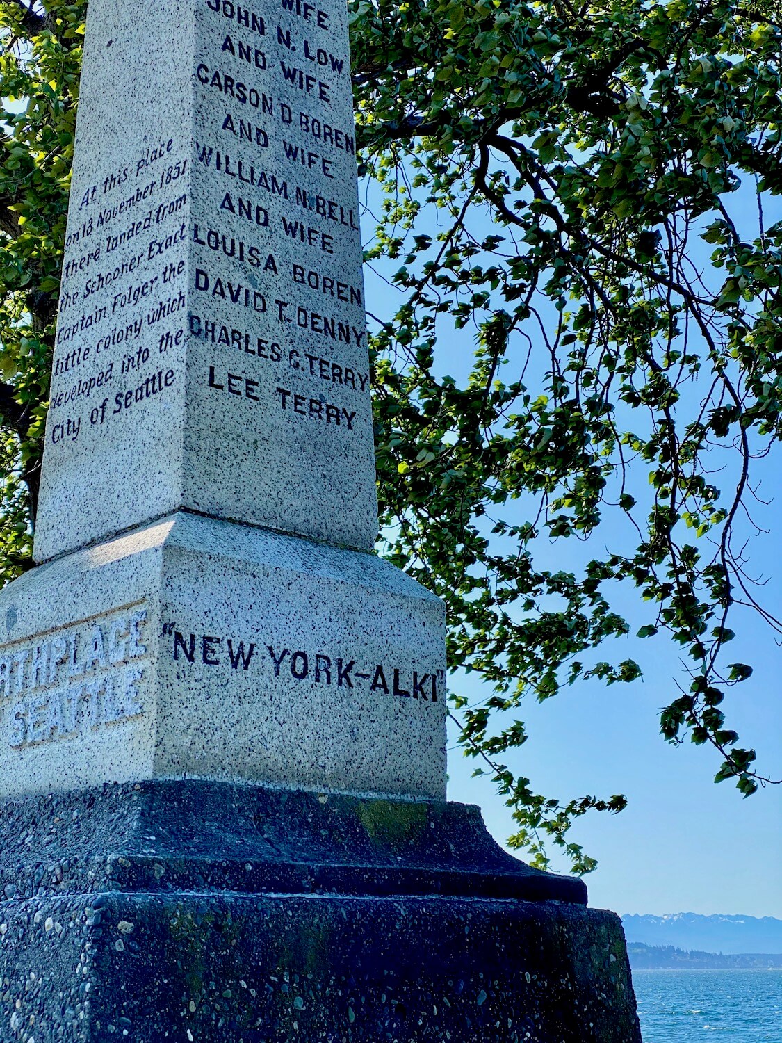 This monument commemorates the birthplace of Seattle and is a granite obelisk shaped tower with black engraved letters that describe how the settlers arrived on this land in 1851. There are leaves of a tree in the background along with the rolling blue waters of the Puget Sound. This is on Alki Beach in West Seattle.