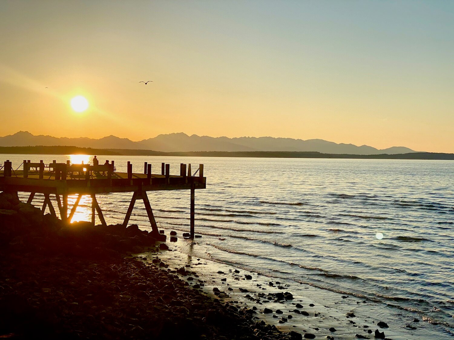 Beautiful spring sunset from Alki Beach in West Seattle. The light waves wash up onto a rocky beach while the pier juts out into the water. The Olympic Mountains in the distance create a backdrop for the peach, yellow and blue colors of the sky with a bright yellow ball of sun getting ready to set.