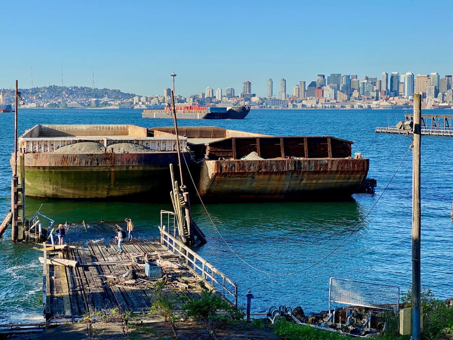 Crossing from Jack Block part to Joe Block Park in West Seattle, a view of the Seattle skyline is visible in the background while two large barges are docked directly in front of a dilapidated railroad dock ending in the Puget Sound. There are three people investigating near the waters edge and the greenish blue sound is flat and calm around the marine vessels.