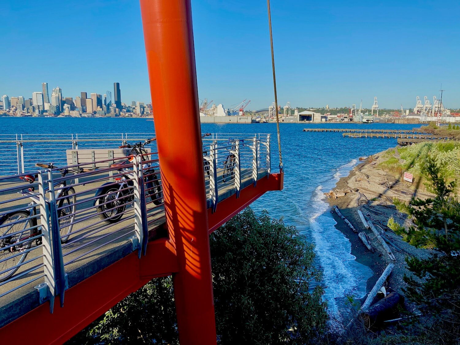 A view from high up in the observation tower at Joe Block Park in West Seattle. The structure is supported by giant orangish red tubes of metal and thick cables. The platform offers sweeping Lewis of the downtown skyline of Seattle, including the tall, black colored Columbia Center in the background. Closer to the tower there are foamy white waves rolling onto a small spit of beach with pieces of driftwood, pebbly sand and low cover bushes. The industrial area of Seattle is directly behind the beach scene.