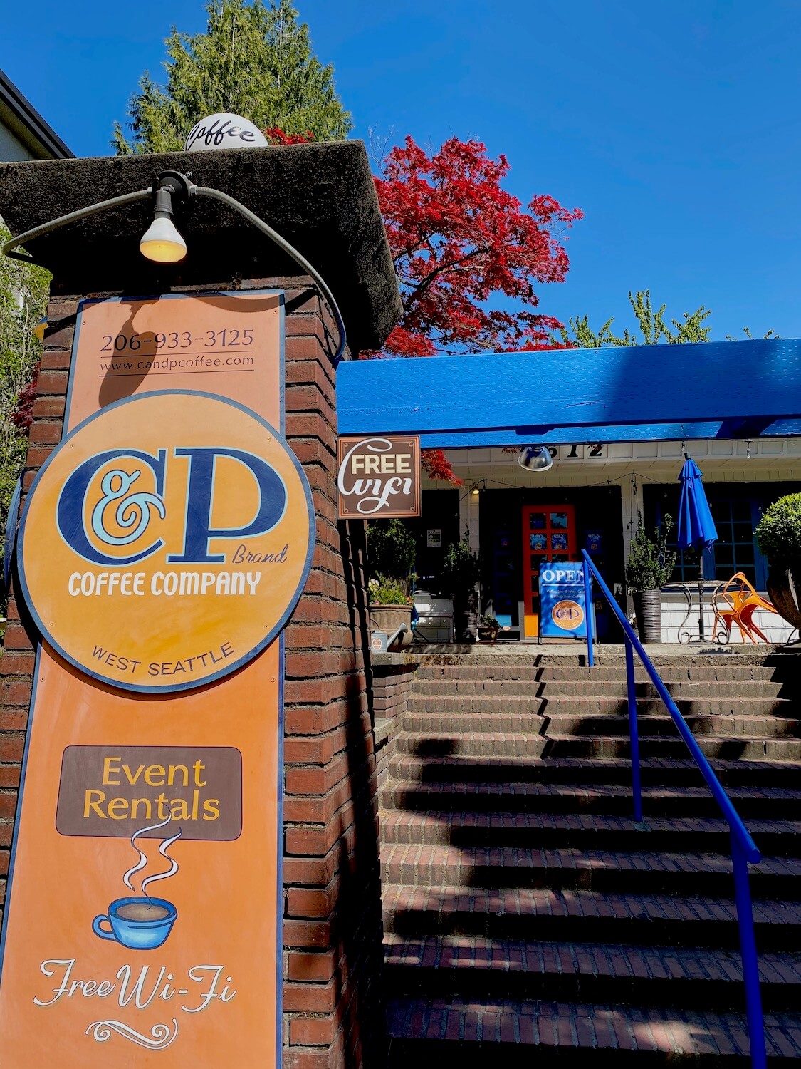 The steps walking up to CP Coffee in West Seattle pass by a large brick entry pillar with an orange sign. In the background there is a bright blue awning and tiny window paned door with orange trim. A pine tree and bright red maple light up the sky, along with the bright blue.