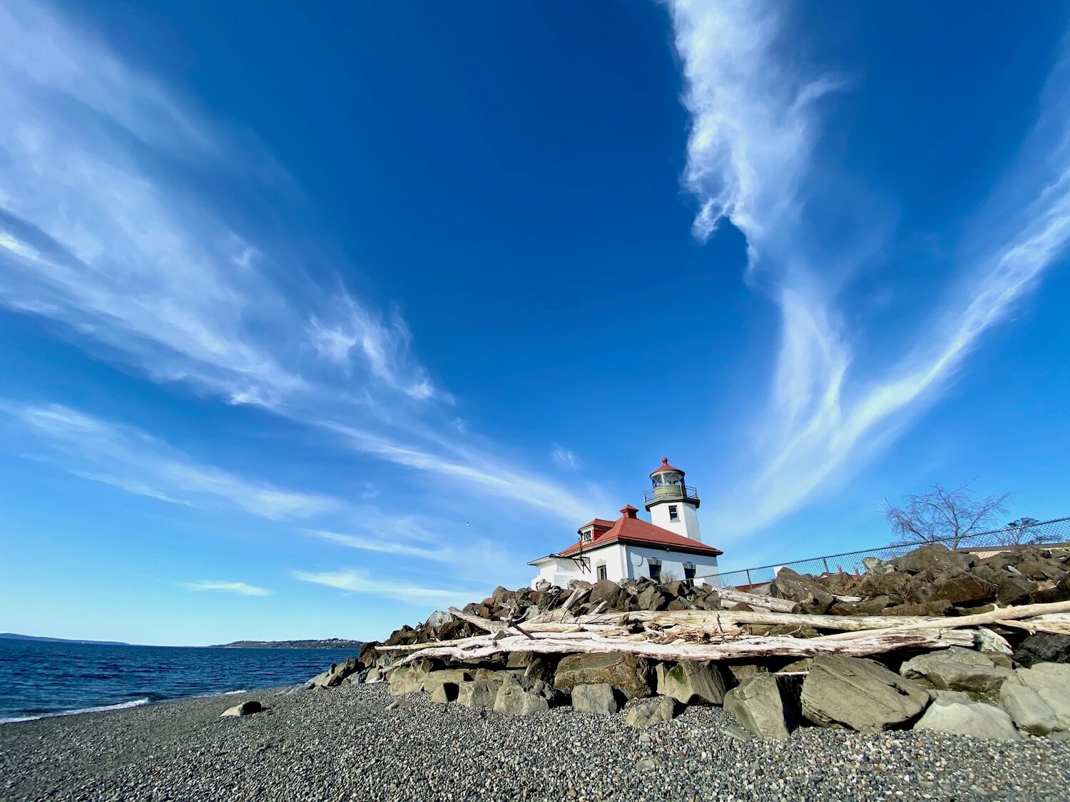 Alki Point Lighthouse is a great thing to do while visiting West Seattle. The pebbles of the beach lead up to larger boulders covered with driftwood. Beyond this seawall is the lighthouse with red tile roof and sharp white colored walls. The sky is blue with streaks of puffy white clouds.