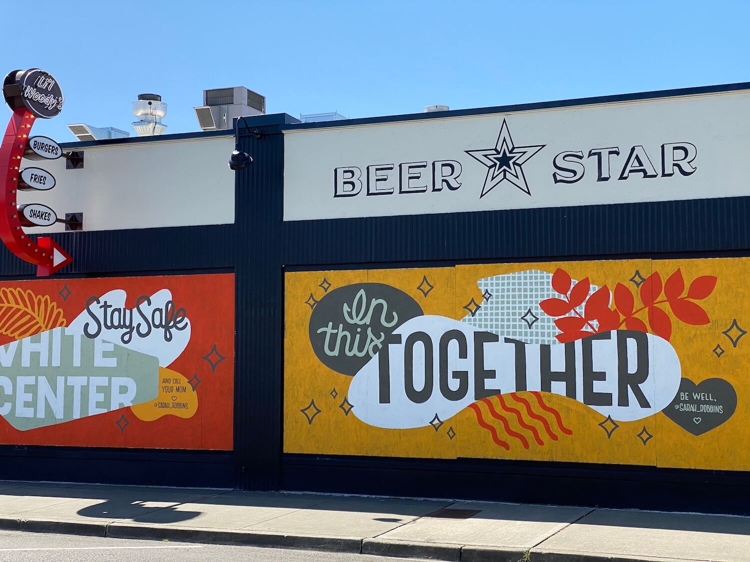 Two building murals in White Center neighborhood in Seattle say, "Stay Safe White Center" and "In This Together." The gray sidewalk sweeps under the art while the bright blue sky illuminates from above.