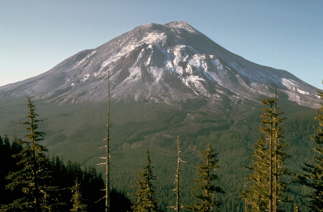 Mt. St. Helens prior to the big eruption on May 17, 1980. The mountain has darkened areas where the glaciers and snow are grayed and rocks are exposed while the lower areas of the mountain are rich with green forest. The sky in the background is blue.
