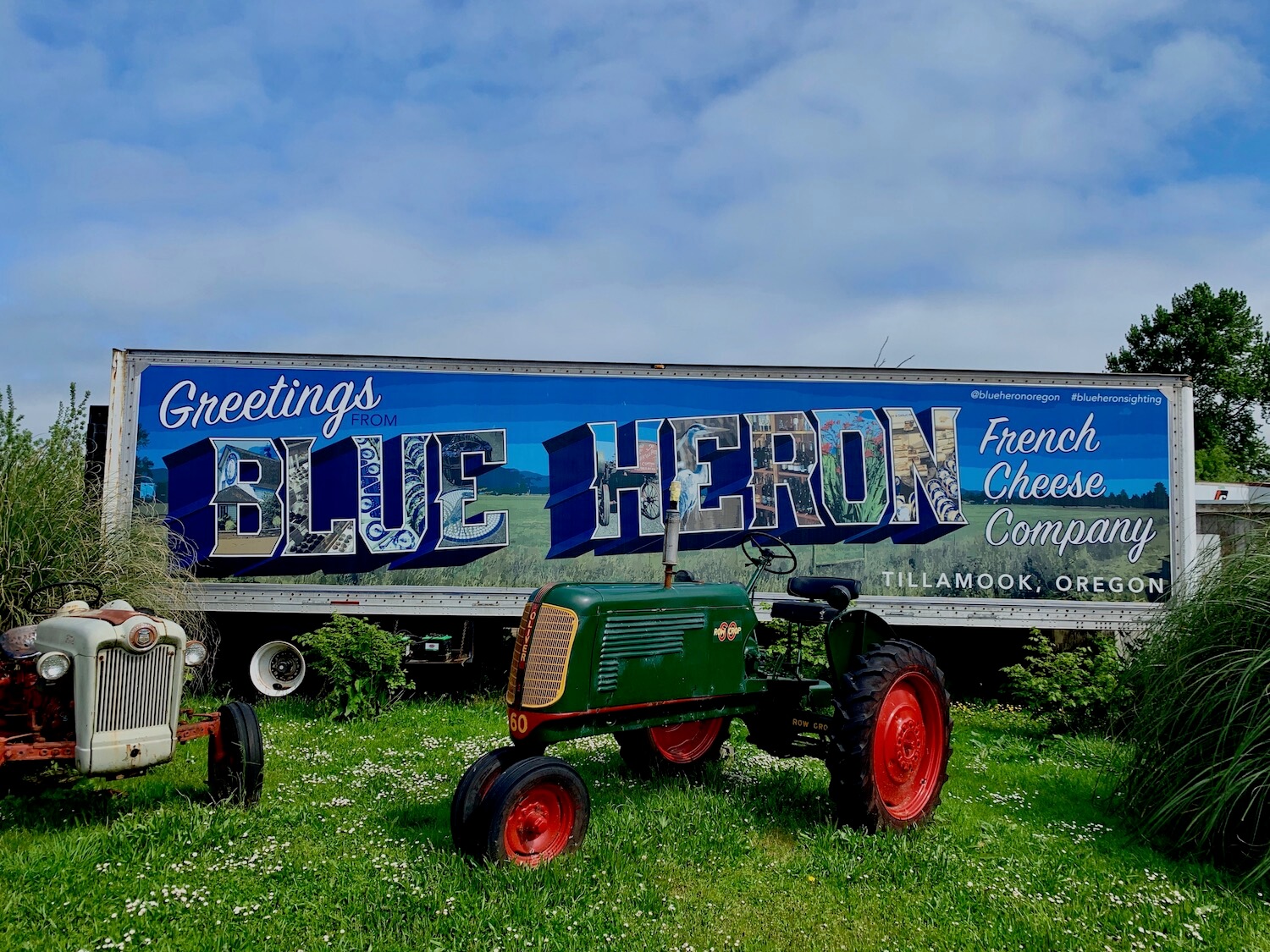 The shot shows a semi-truck trailer painted with an old fashioned post card style to promote Blue Heron French Cheese company in Tillamook, Oregon. In front of the truck, on a patch of green grass sit two retro tractors. One is green with red trim and the other white with red trim. The sky is a bright blue.